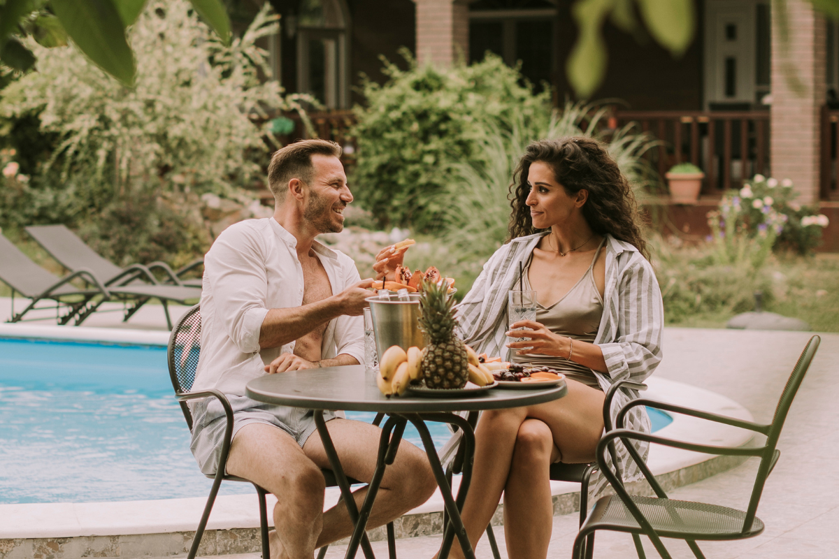 Pareja relajándose junto a la piscina de un hotel, compartiendo fruta y bebidas en una mesa al aire libre.